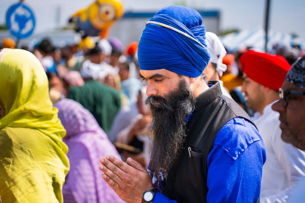 A Sikh man in traditional attire prays at an outdoor gathering, surrounded by a colorful crowd.