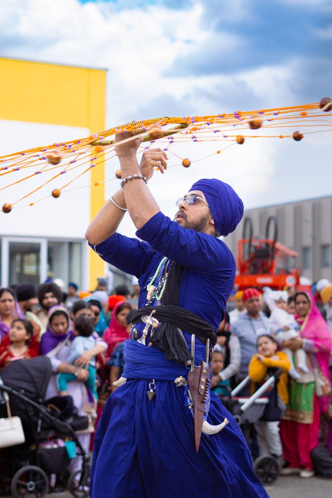 Sikh performer in blue demonstrating traditional martial art at a street event surrounded by a diverse crowd.